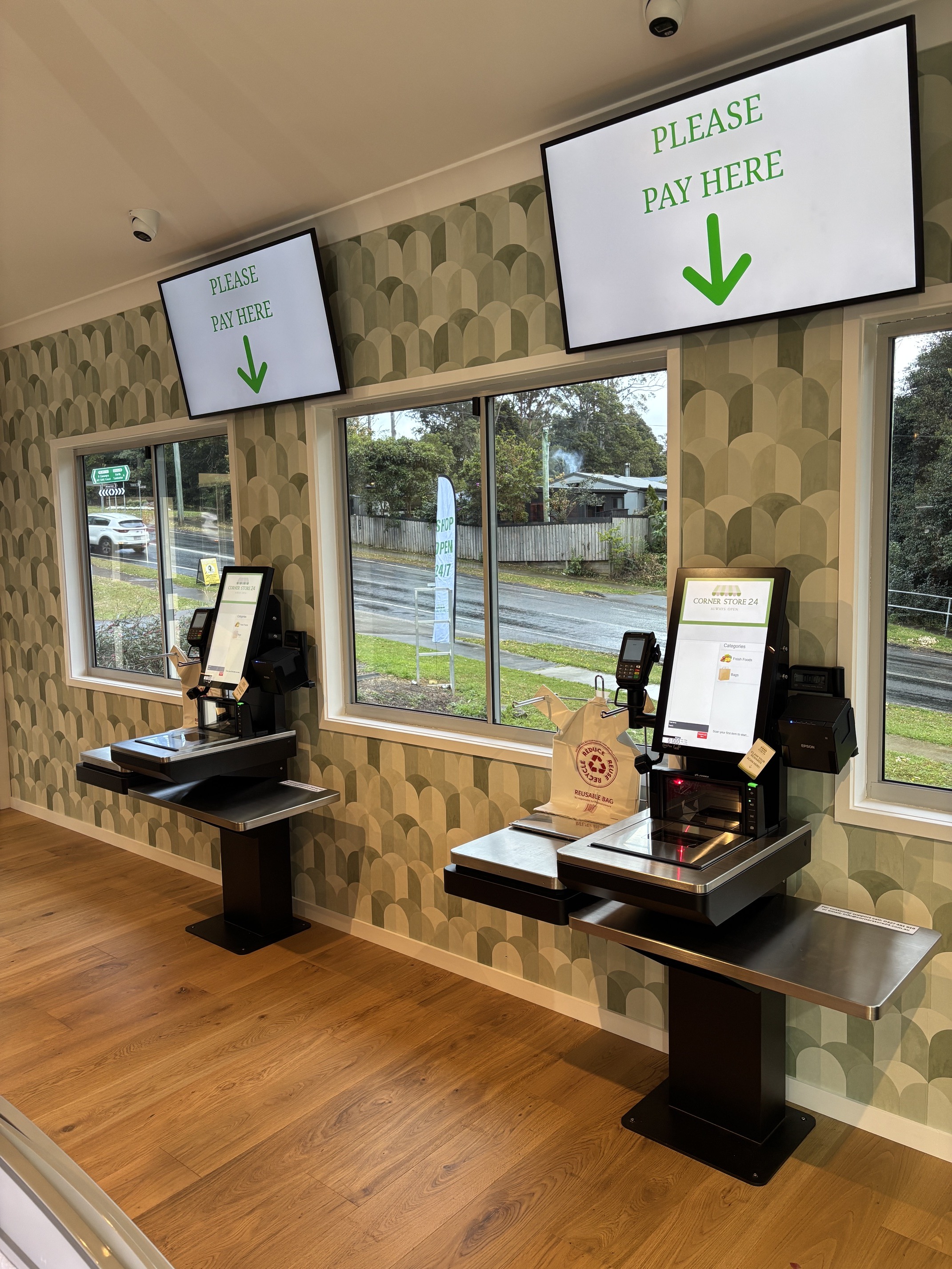 Two self-checkout terminals with scanners, scales and card readers beneath large Please Pay Here screens